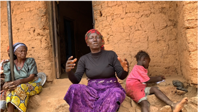 Three people sit outside a mud brick house. A woman gestures, another sits silently, and a child writes on paper.