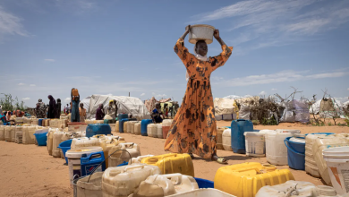 A woman carries a bucket on her head near rows of water containers in a dry landscape with tents and people in the background.
